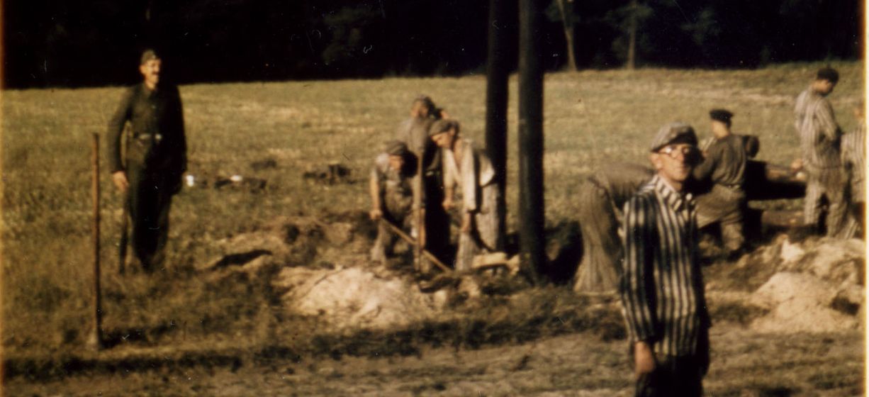 Das Farbbfoto zeigt Häftlinge des KZ-Außenlagers Lieberose in gestreifter Kleidung, die einen Strommast eingegraben haben, sowie SS-Männer in Uniform. Das Foto, von dem hier ein Bildausschnitt zu sehen ist, wurde im August 1944 von einem SS-Mann aufgenommen.
