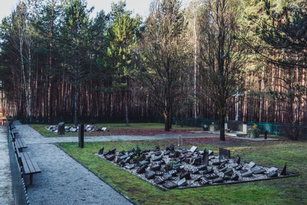 Auf dem Waldfriedhof mit Opfern des sowjetischen Speziallagers befindet sich rechts ein großes Holzkreuz. Auf der Rasenfläche davor sind zwei Felder mit zahlreichen kleinen Steintafeln zu sehen, die einzelnen Verstorbenen gewidmet sind.