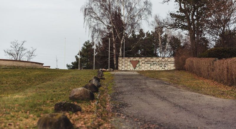 Zugang zum Mahnmal in Lieberose, das sich auf einer Anhöhe befindet, 2025 (Foto: SBG/Peschel) Ein leicht ansteigender Weg führt zum Mahnmal in Lieberose, dessen Gedenkwand mit rotem Dreieck in der Bildmitte zu sehen ist.